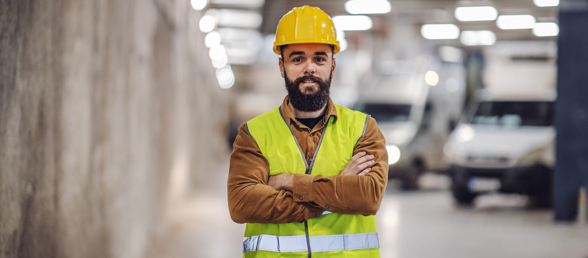 Bearded man in yellow hard hat and vest, crossing arms and smiling